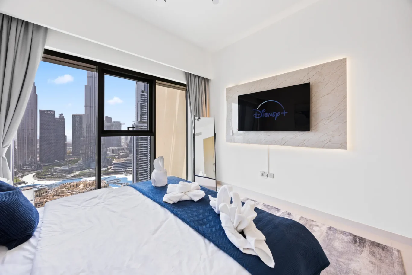 Hotel room with a large window view of a city skyline, bed with white linens and a blue runner, towel swans on the bed.