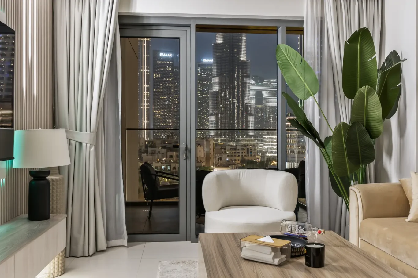 City skyline at night visible through glass balcony doors in a modern living room with a white curved chair, beige sofa, and a tall indoor plant.