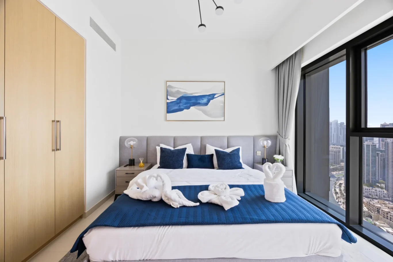 Hotel bedroom with large windows showing a city skyline; bed has blue accents and towel swans on the duvet.