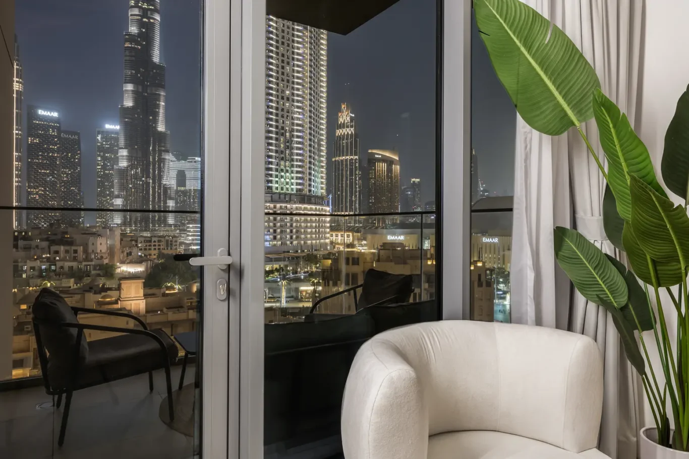 Nighttime city view through a glass balcony door; a modern living room with a cream curved chair nearby and a tall green plant in the foreground.
