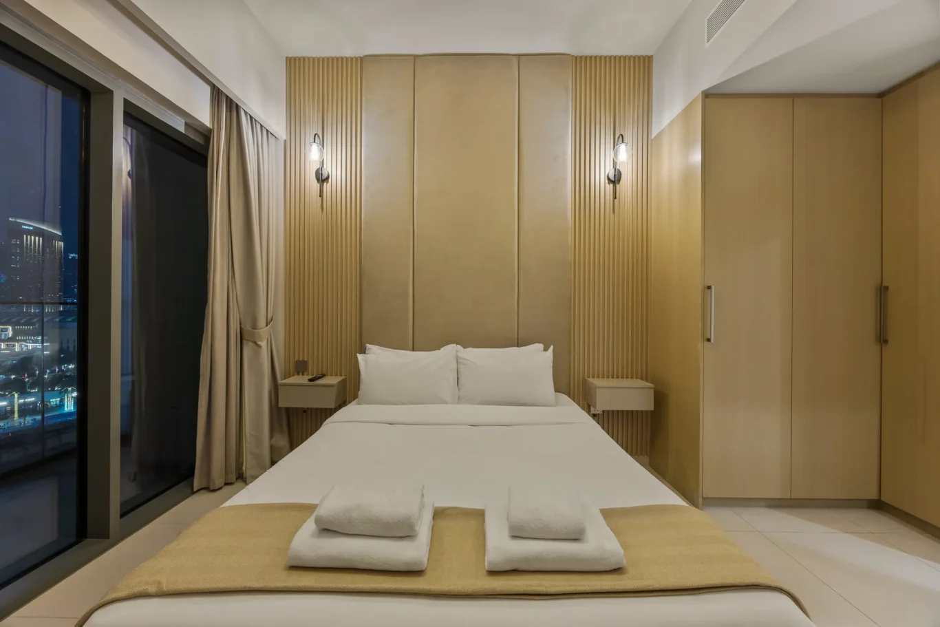 Modern hotel bedroom featuring a double bed with white linens, beige headboard panels, wall sconces, and matching nightstands; large window with curtains shows city lights outside at night.