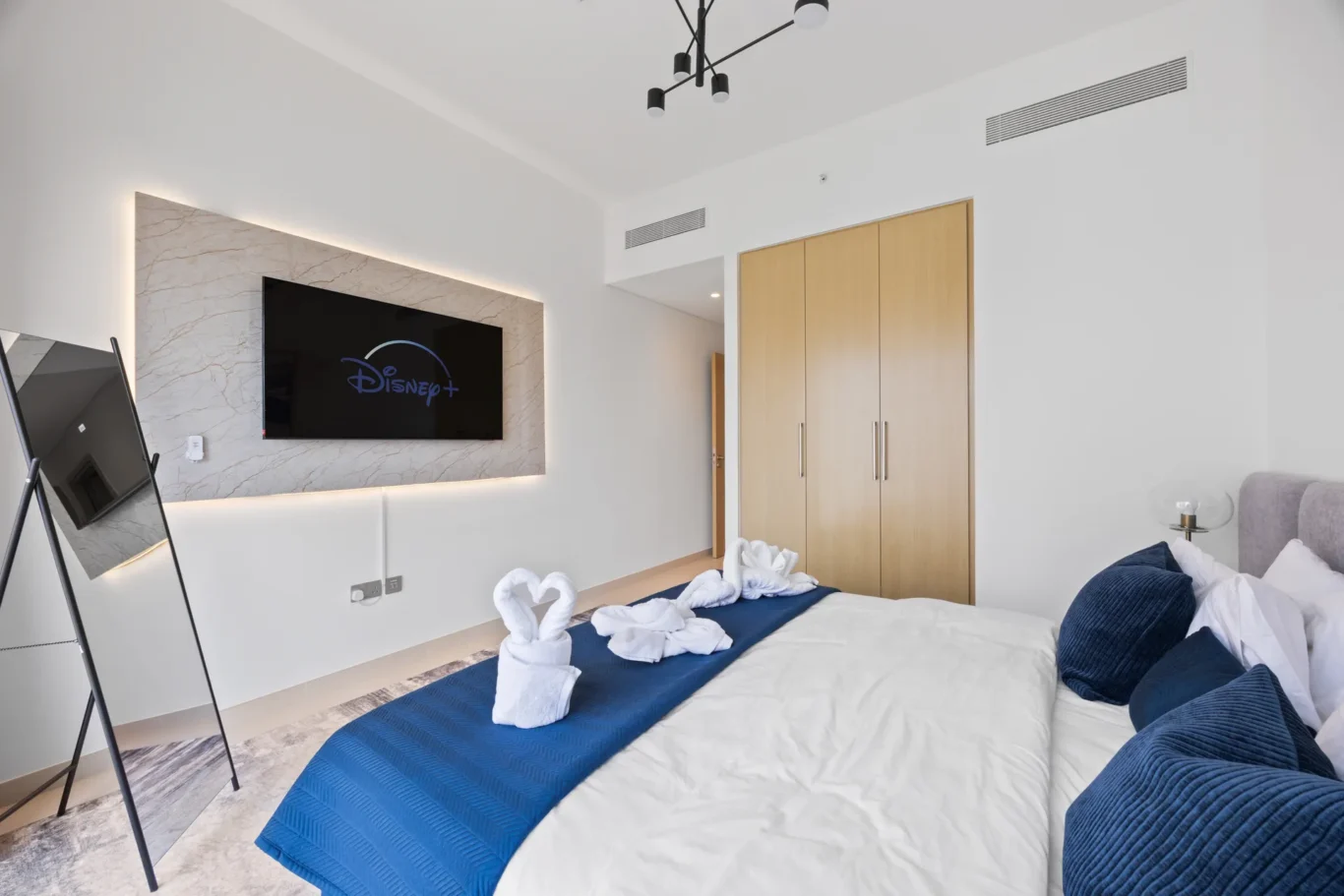Modern hotel room with two twin beds, blue bed runners, towel swans, and a wall-mounted TV on a marble panel beneath ambient lighting.
