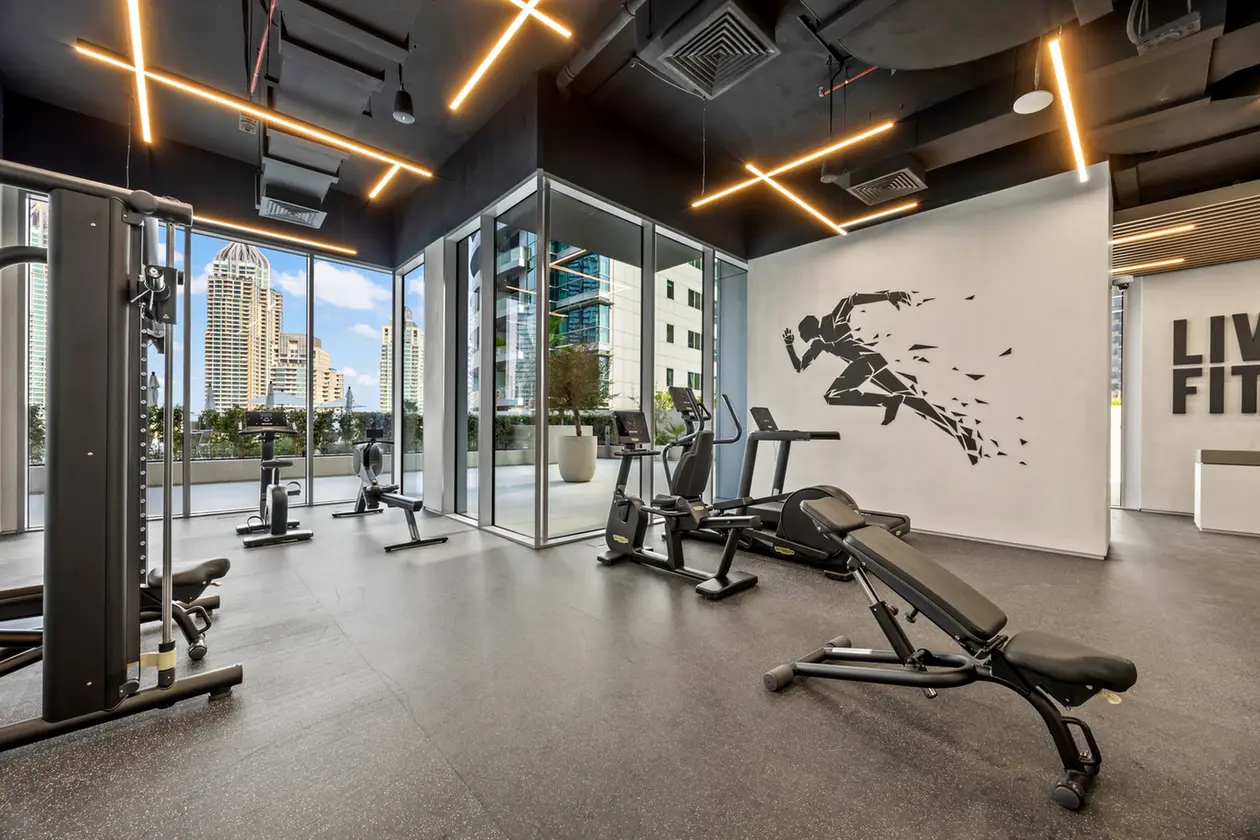 Modern gym with treadmills, stationary bikes, and weight machines near large windows overlooking city skyscrapers at dusk/daylight.