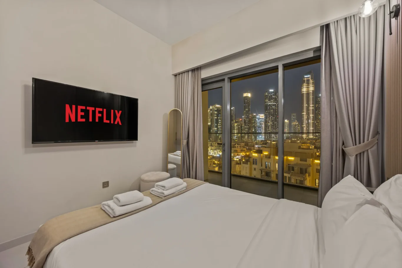 Hotel room at night with a wall-mounted Netflix screen, two neatly made beds, and folded towels on a beige runner at the foot of the beds.