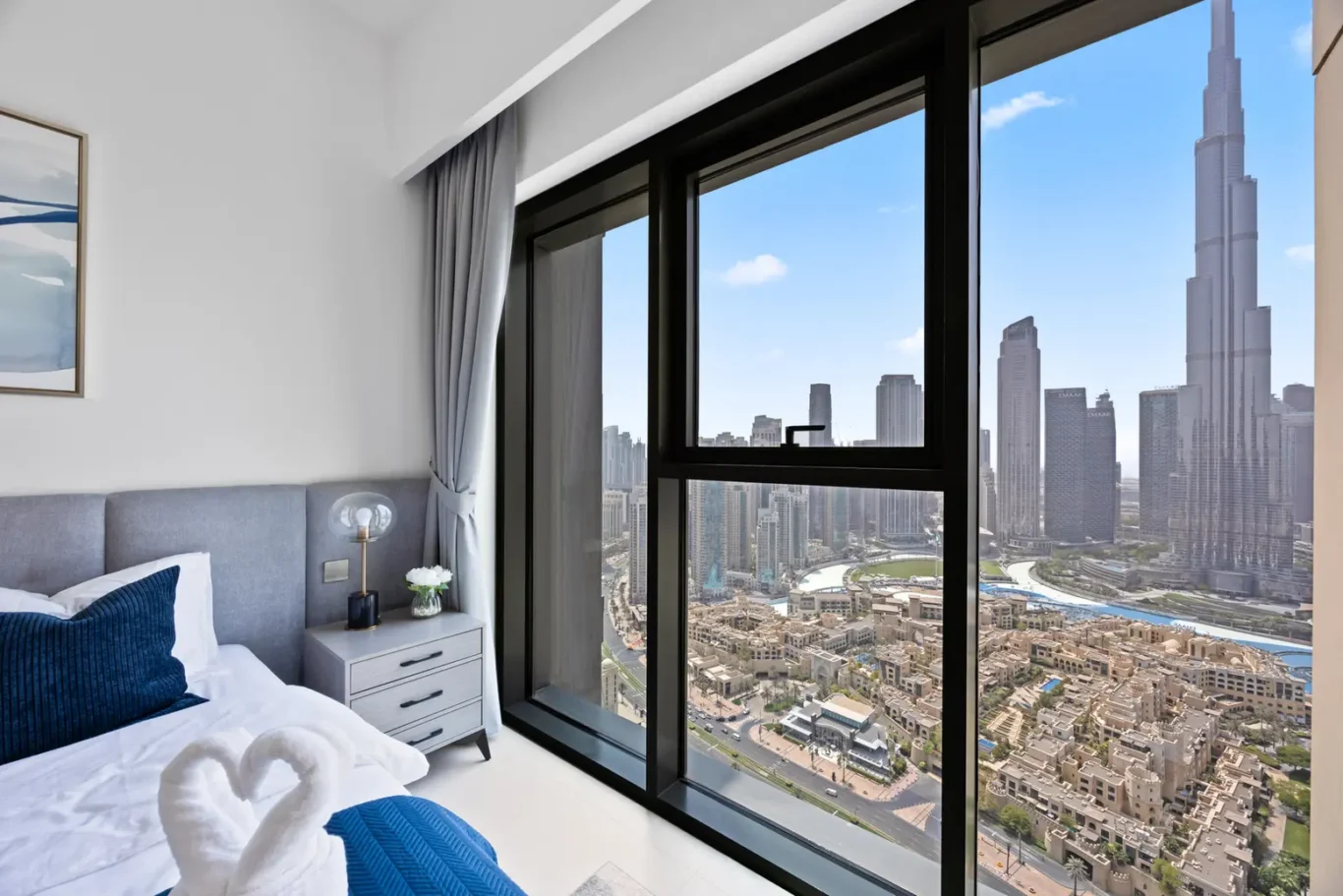 Bright hotel bedroom with a large window showing a city skyline and tall skyscrapers; bed with blue pillows and a nightstand nearby.