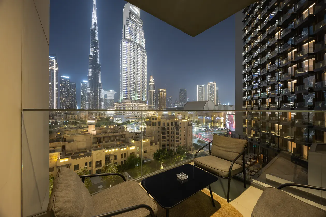 Nighttime city skyline viewed from a balcony with glass railing, four cushioned chairs, and a small glass table overlook lit skyscrapers below for an urban living vibe.