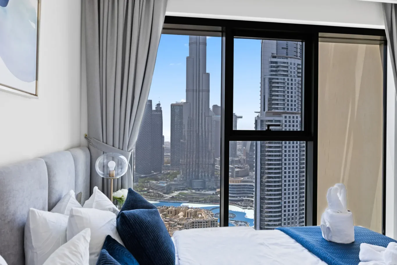 Luxurious hotel bedroom with white bedding, blue cushions, and a cityscape view of tall skyscrapers through a large window.