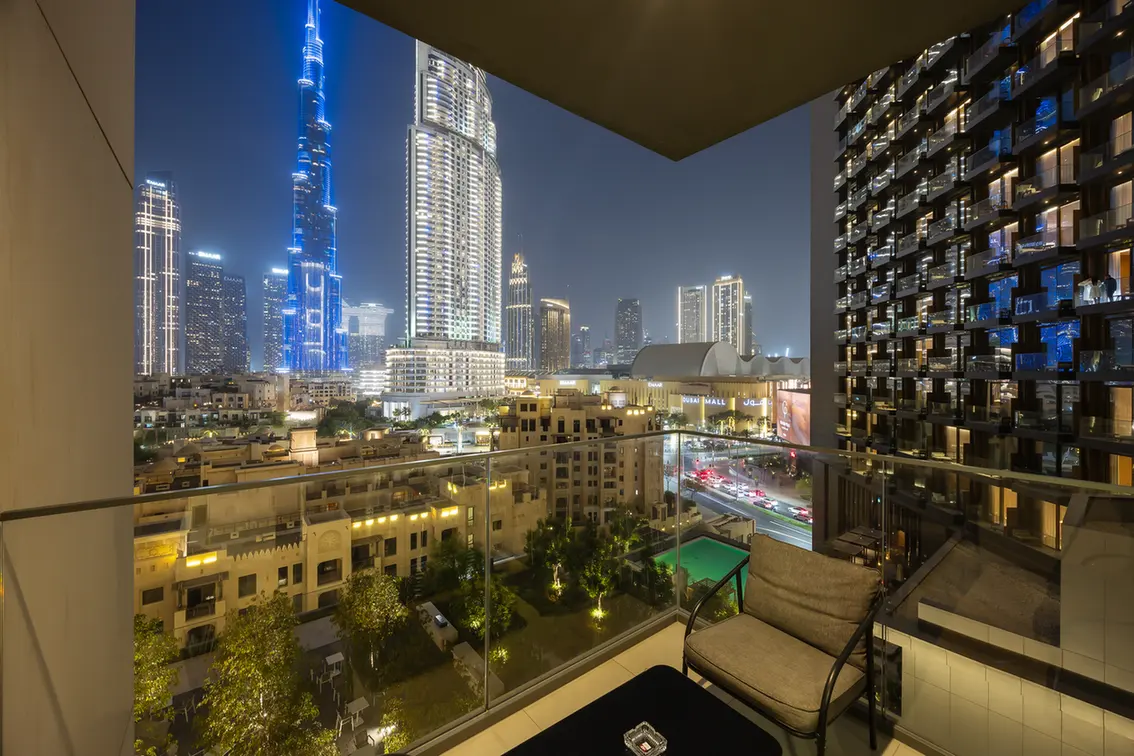 Nighttime city skyline seen from a balcony with glass railing and outdoor seating. Tall, illuminated skyscrapers dominate the view.