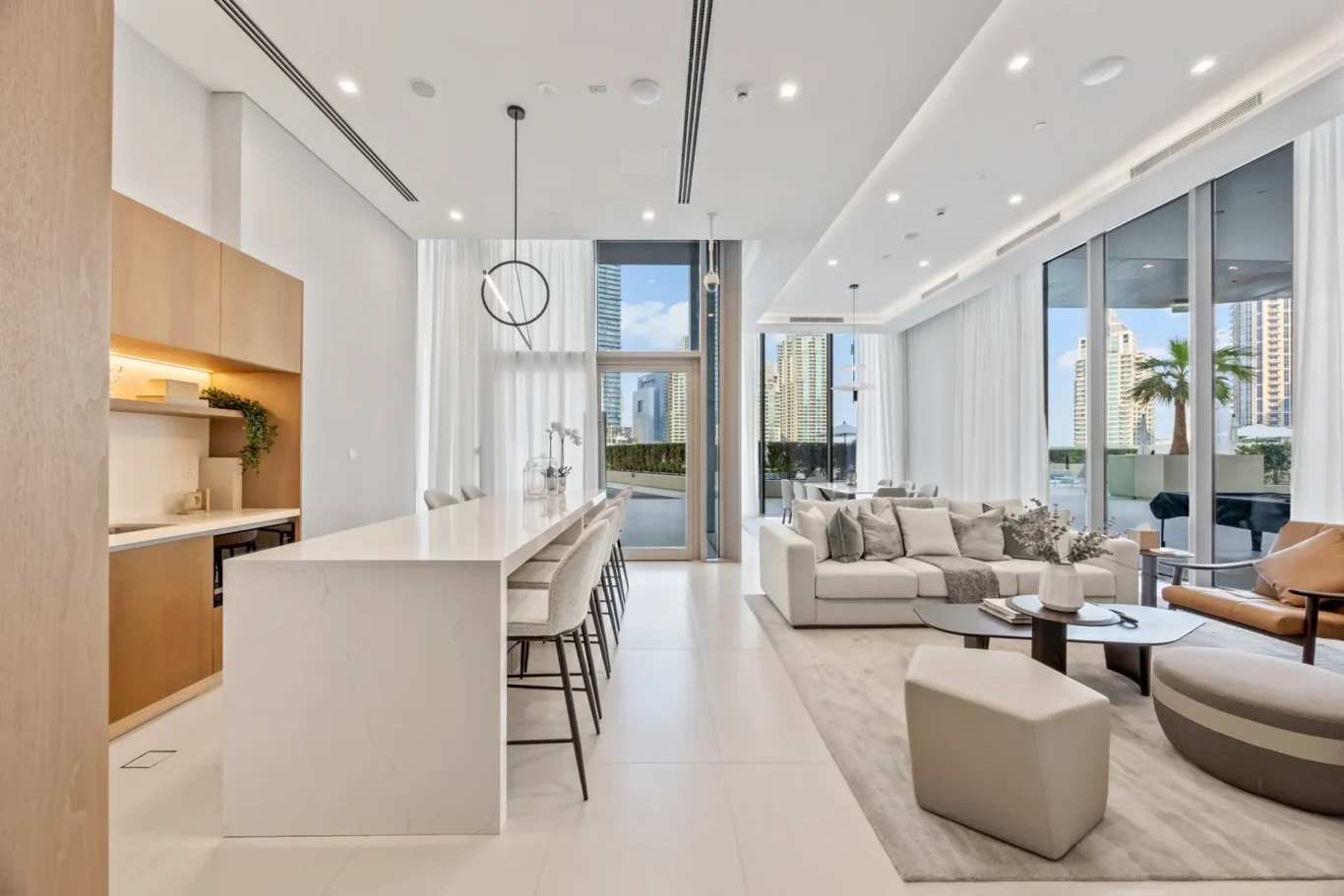 Open-concept living area with white kitchen island, beige cabinetry, and a bright lounge overlooking city skyline through floor-to-ceiling windows.