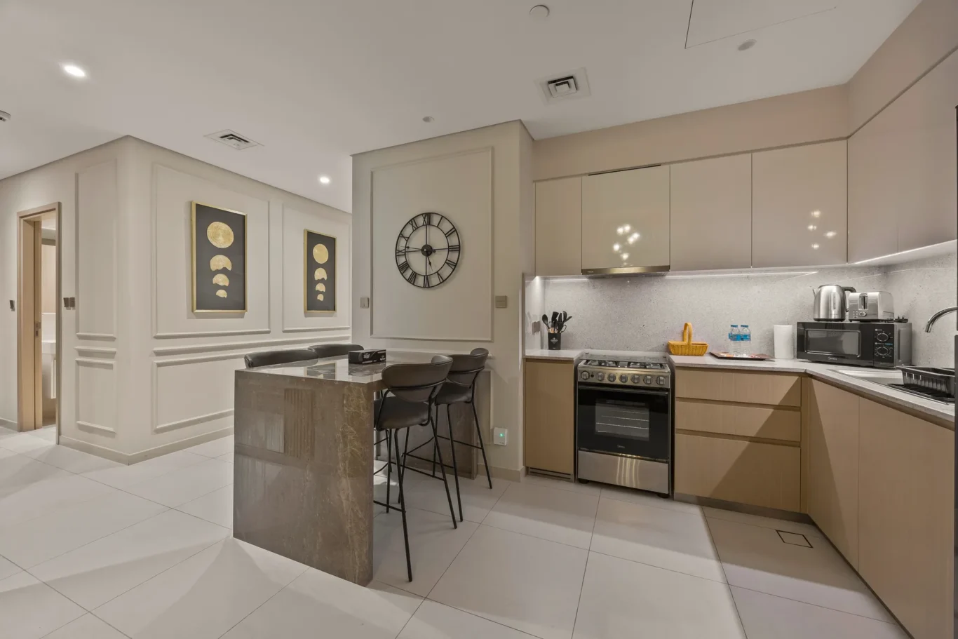 Open-plan kitchen with an island and four bar stools, beige cabinets, and a wall clock beside framed moon-phase art on paneled walls of the dining area.