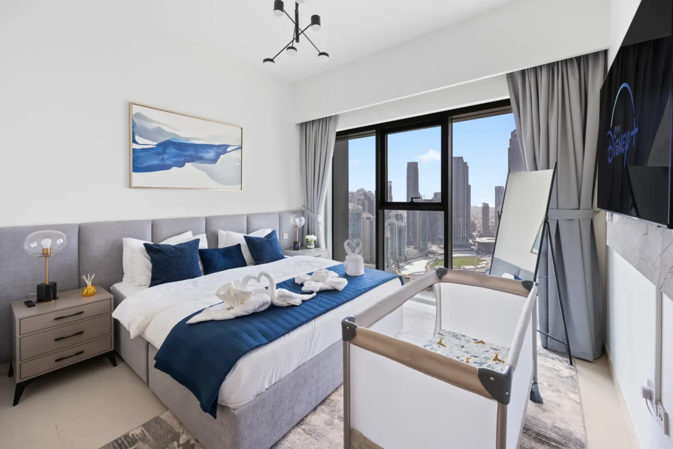 Modern hotel bedroom with a gray upholstered bed, blue bedding, and city skyline visible through a large window; a crib is at the foot of the bed.