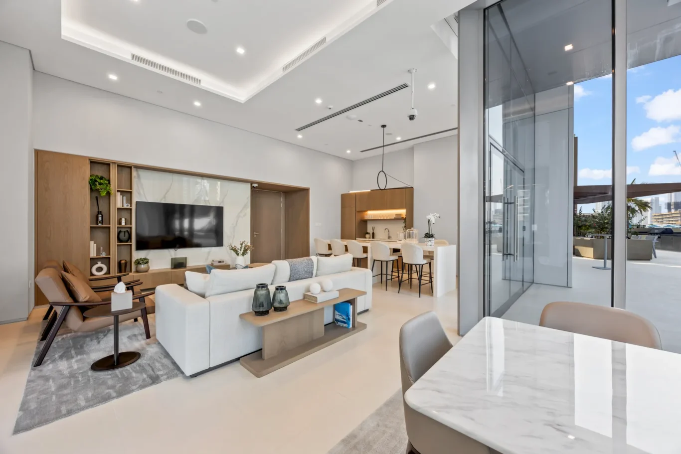 Spacious modern living room with white sofas, wooden shelves, and a wall-mounted TV, adjacent to a kitchen and dining area.