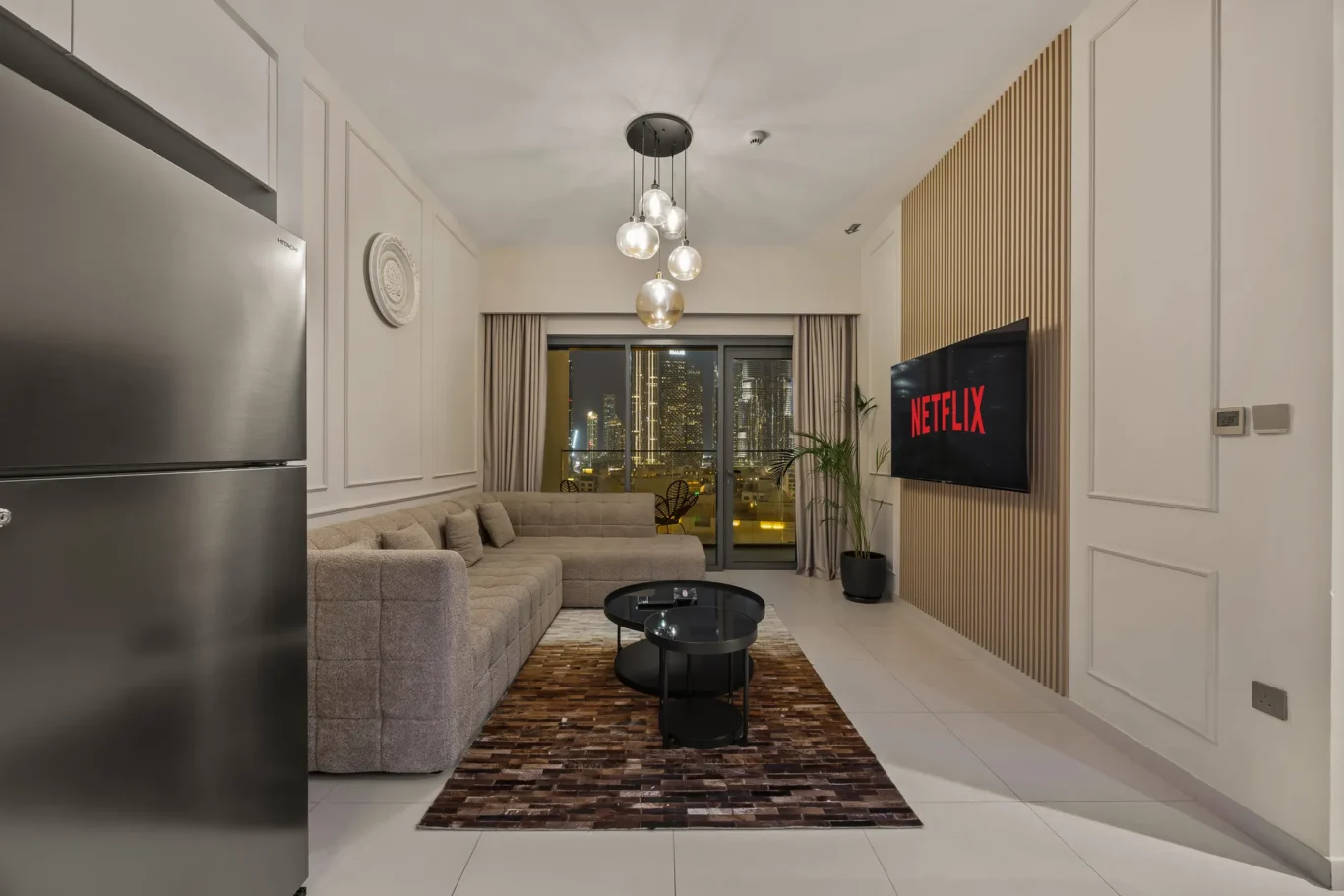 Living room with a beige sectional sofa, glass coffee tables, and a wall-mounted Netflix screen; city lights seen through the balcony window at night.