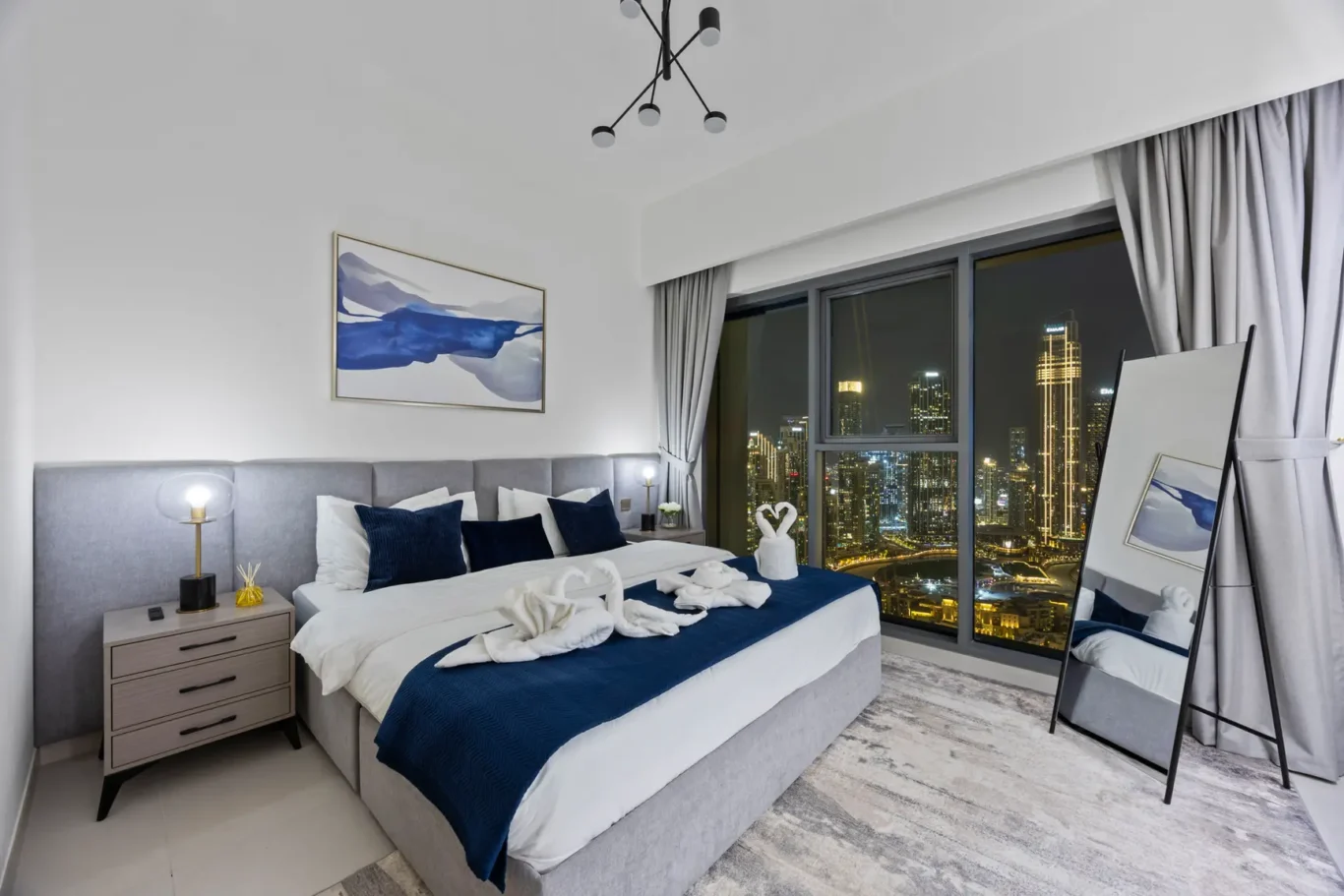 Hotel bedroom with gray upholstered bed, white linens, navy accents, and city lights visible through large windows at night.