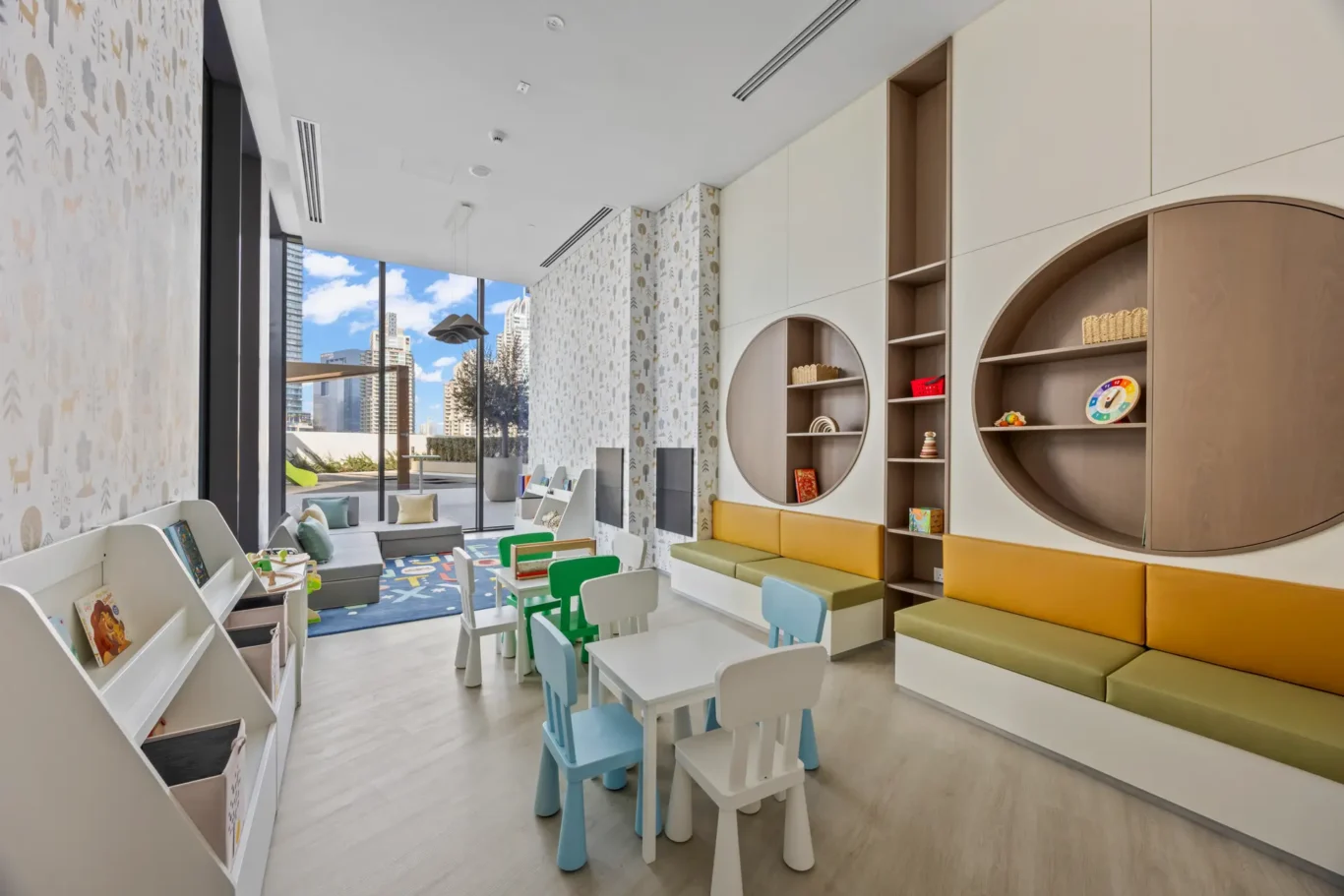 Bright children's playroom with small white tables and pastel chairs, circular wall shelves, and large windows showing a cityscape.