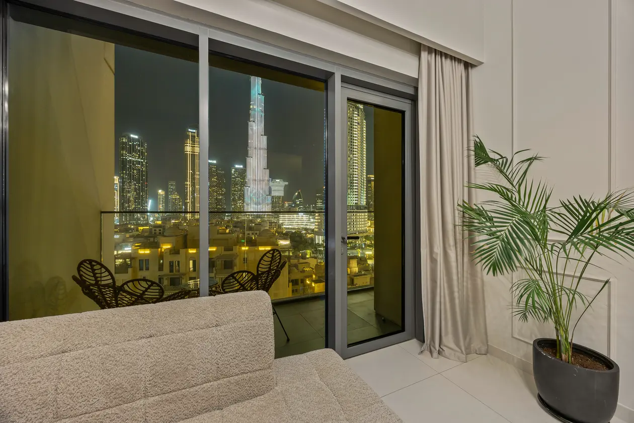 Nighttime city skyline seen through large glass doors; beige couch in foreground and a tall potted plant on the right inside the room.