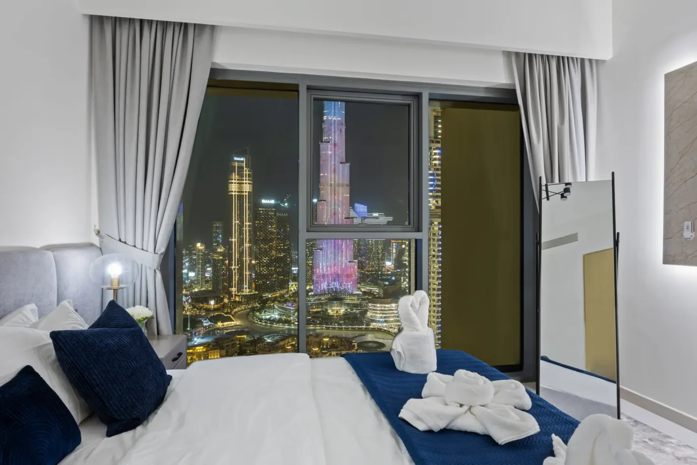 Modern hotel room at night with a large window showing a brightly lit city skyline; bed with white linens, navy pillows, and a blue throw.