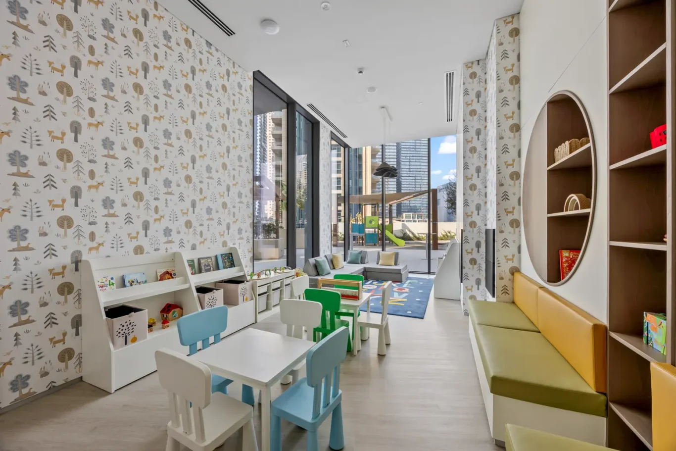 Bright kids' playroom with white furniture, pastel chairs, and bookshelves; nature-themed wallpaper and large windows overlooking the city.