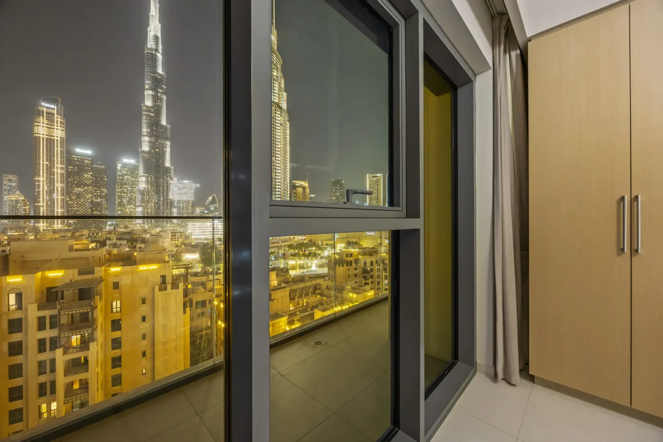 Nighttime city skyline visible through a large glass door of a modern apartment, with a balcony and light wood cabinets on the right.