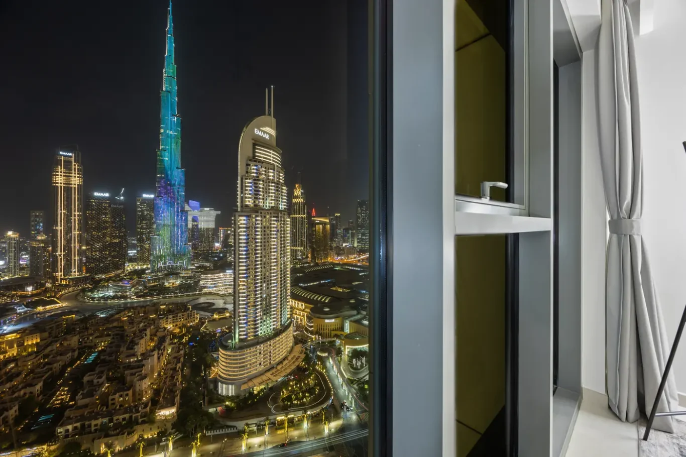 Nighttime city skyline seen from a modern high-rise window, with illuminated skyscrapers and a curved blue-lit tower outside.