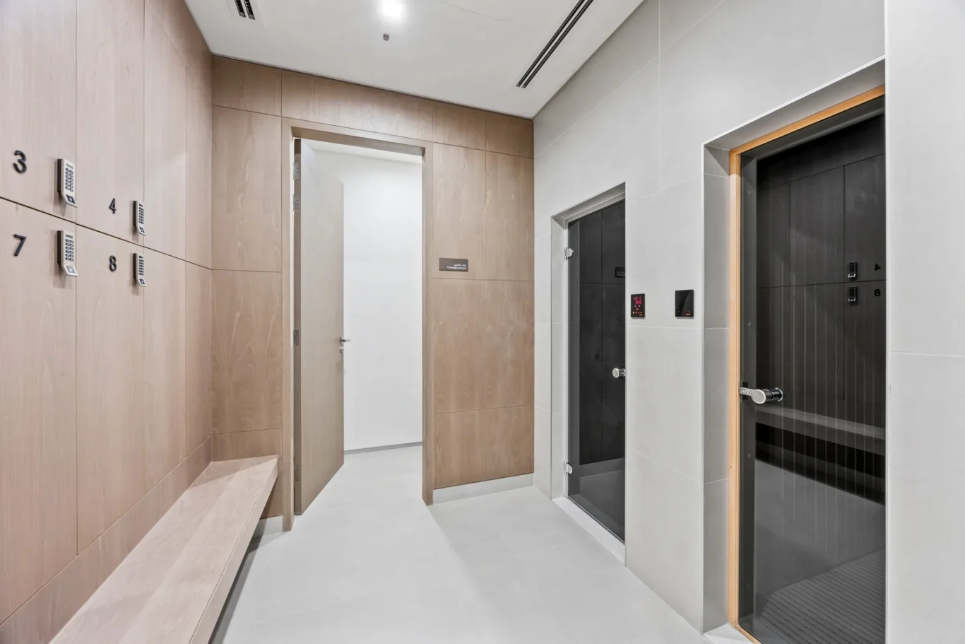 Modern elevator lobby with light wood wall panels, a long bench along the left, wall-mounted numeric safes, and a glass-front elevator on the right.