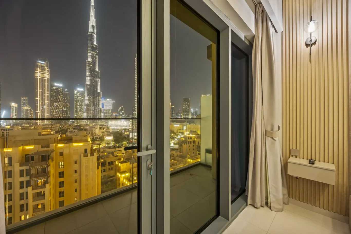 Night city skyline seen from a high-rise balcony with glass doors, beige curtains, and a wooden panel wall inside the room at night.