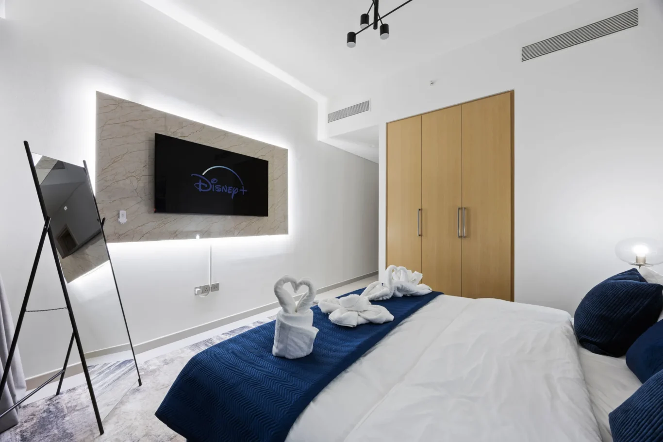 Hotel bedroom with white walls, a wall-mounted TV on a marble panel, a blue bed runner, and folded towel swans on the bed.