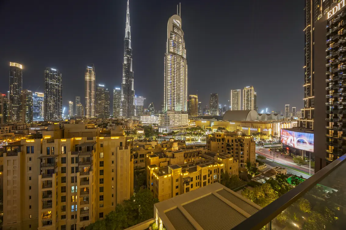 Nighttime cityscape of Dubai with illuminated skyscrapers, including the Burj Khalifa, viewed from a balcony over warm, yellow-lit buildings in the foreground.