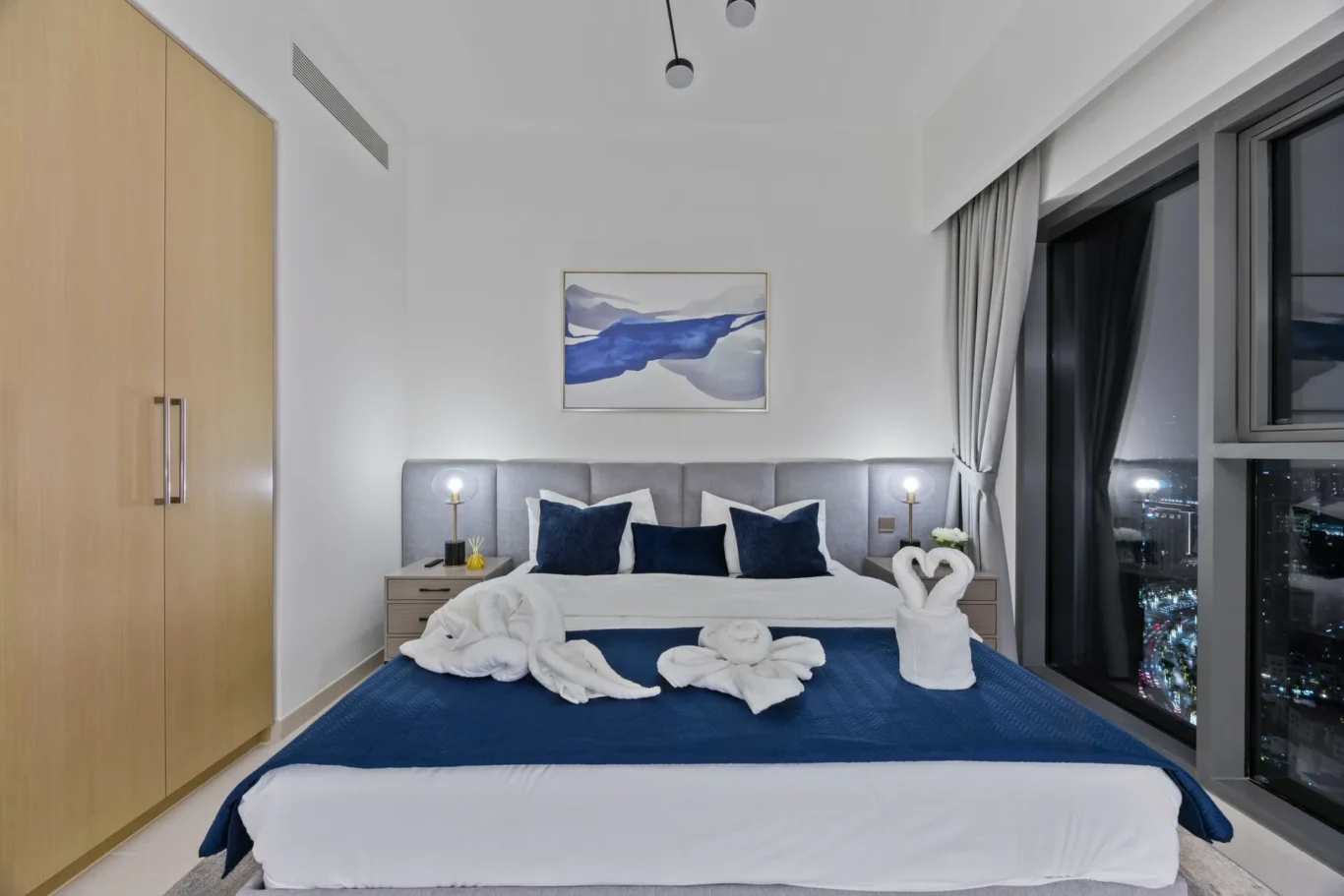 Hotel bedroom featuring a large bed with white linens, blue accent pillows, and towel swans on the bed; city lights visible through floor-to-ceiling windows.