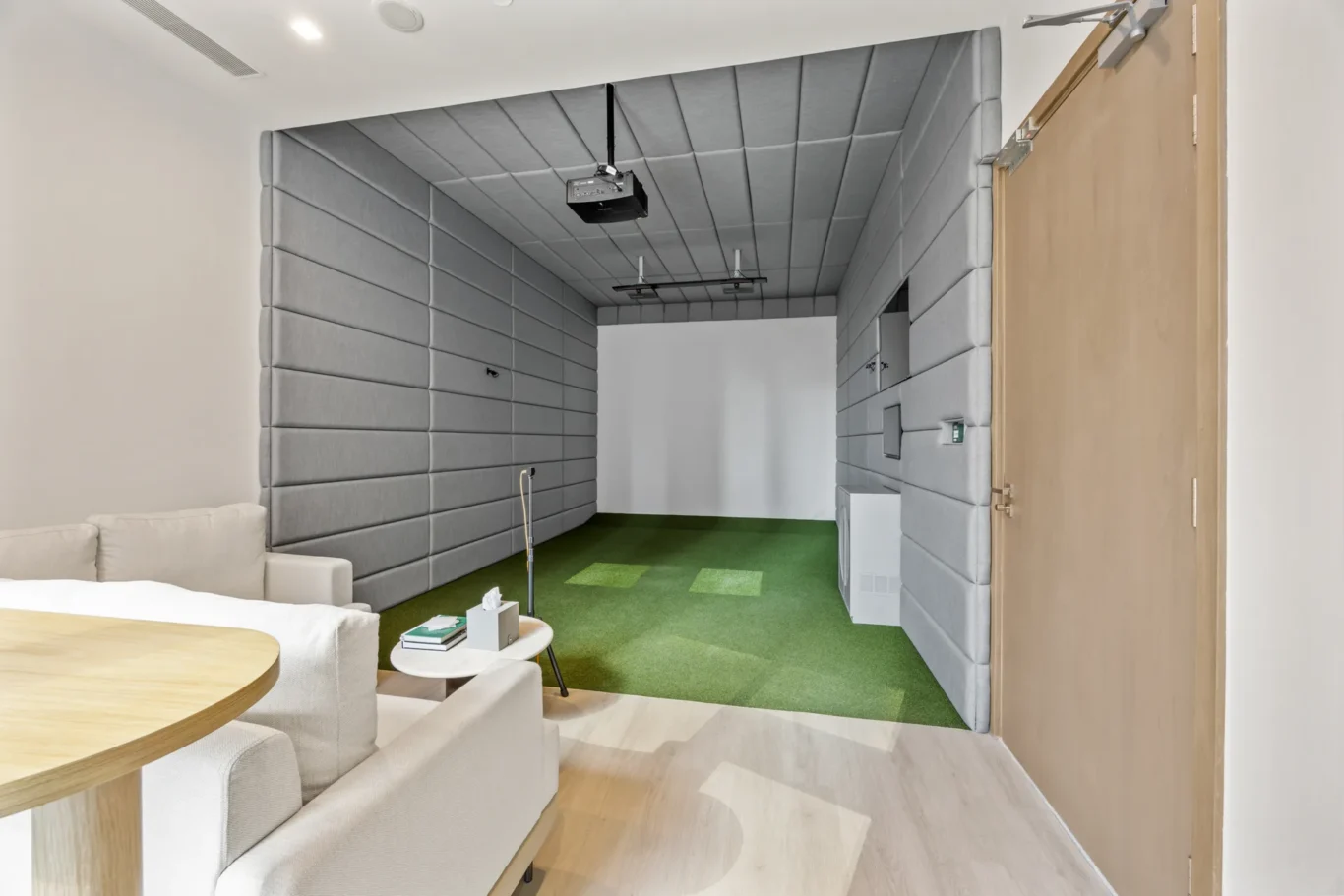 Soundproof meeting room with cushioned gray walls, a ceiling projector, and a green carpet, seen from a seating area with a round wooden table in the foreground