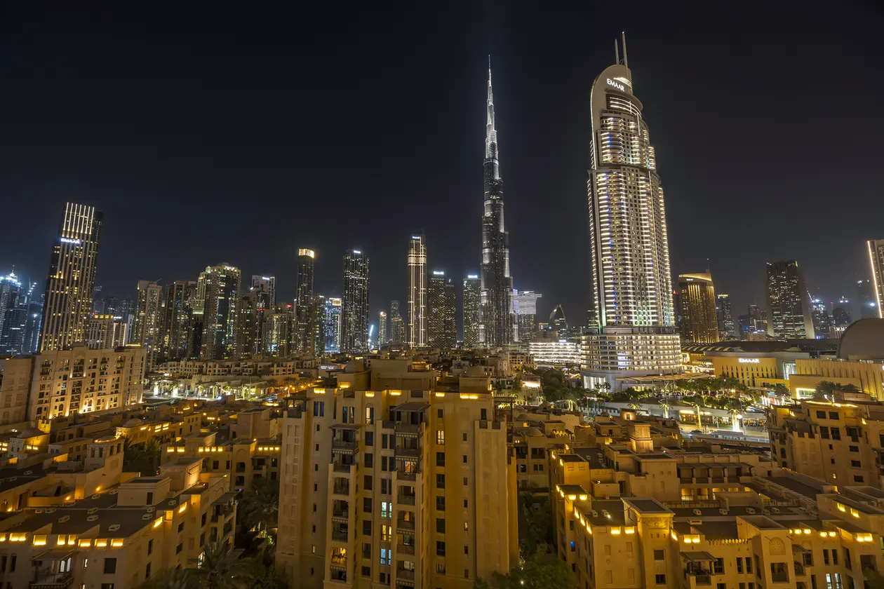 Night view of Dubai skyline with Burj Khalifa towering among illuminated skyscrapers and warm residential buildings in the foreground.