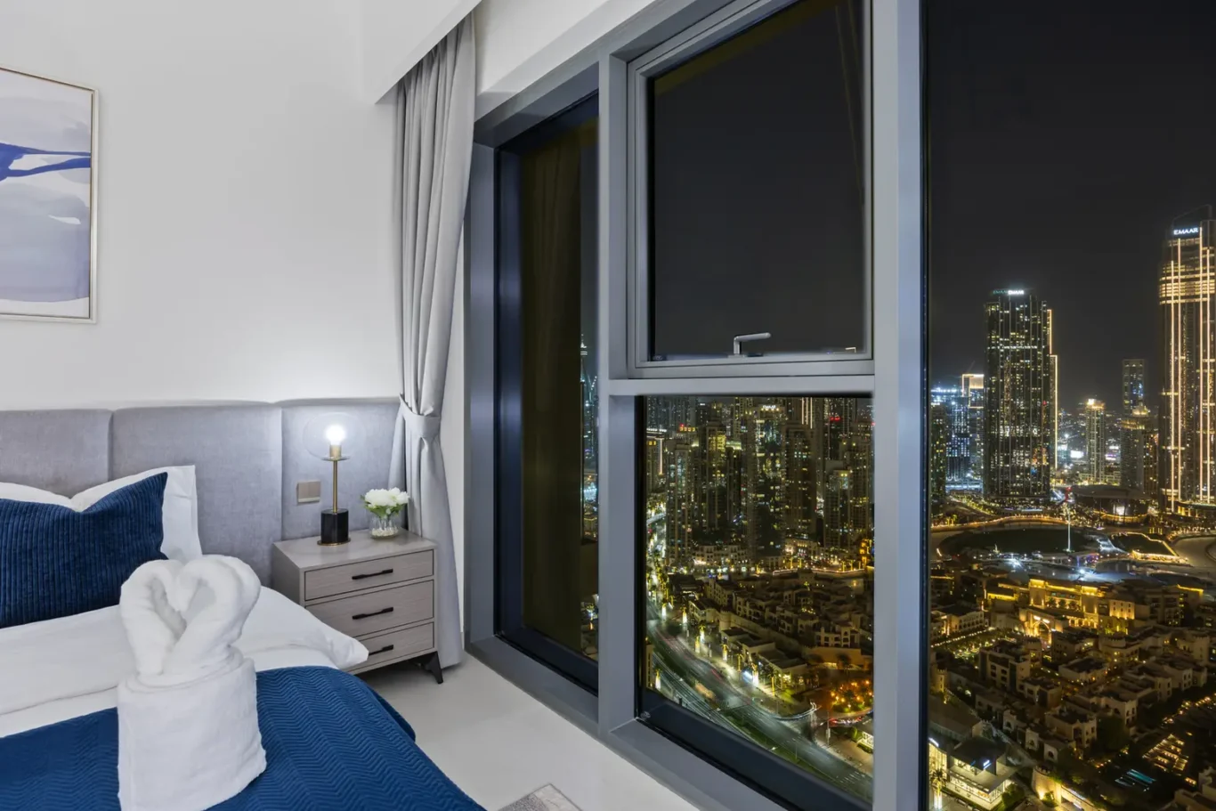 Modern hotel room at night with a city view through a large window; bed with blue bedding and folded towel art on top, plus a nightstand and lamp.