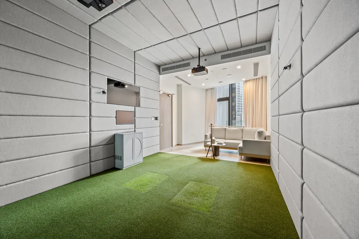 Soundproof room with padded gray walls and green carpet; doorway leads to a living area with sofa and desk near a window.