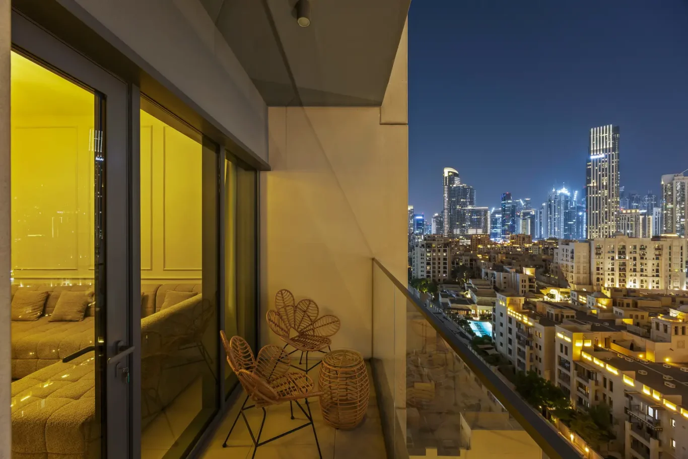 Night-time view from a high-rise balcony: wicker chairs and a lantern on the patio, city skyline lit up in the distance.