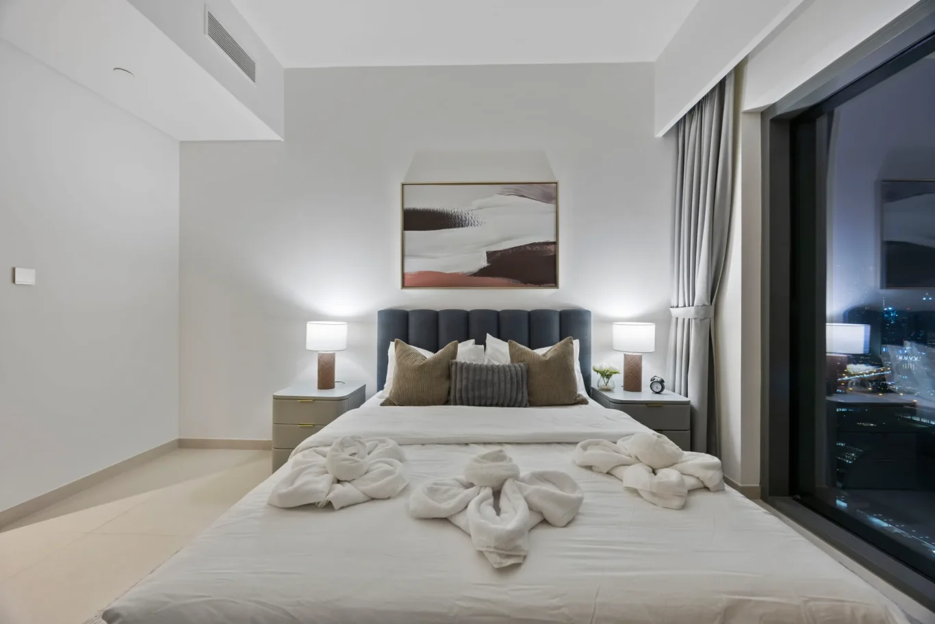 Hotel bedroom with a large bed, gray headboard, matching pillows, two nightstands with lamps, and an abstract painting above the bed; city lights visible through a floor-to-ceiling window at night.