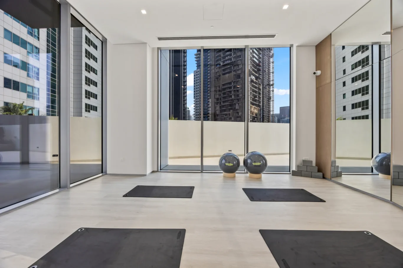Indoor fitness studio with black yoga mats on a light floor, large glass walls, and a city skyline visible outside.