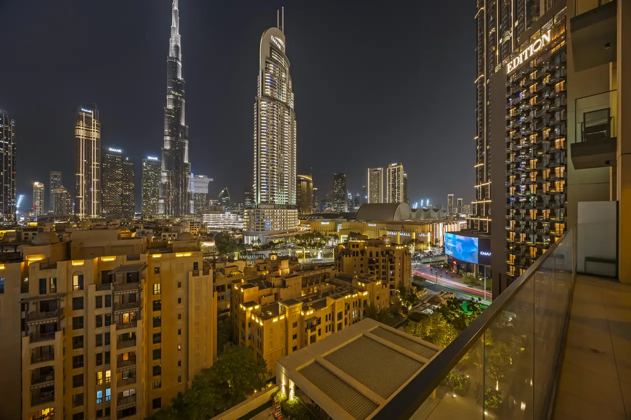 Nighttime cityscape of a modern skyline with tall, illuminated skyscrapers viewed from a balcony railing at the foreground (Dubai-like scene).