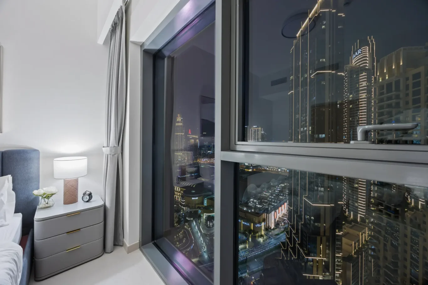 Hotel bedroom corner at night with large windows revealing a lit city skyline; bedside table, lamp, and curtains nearby.