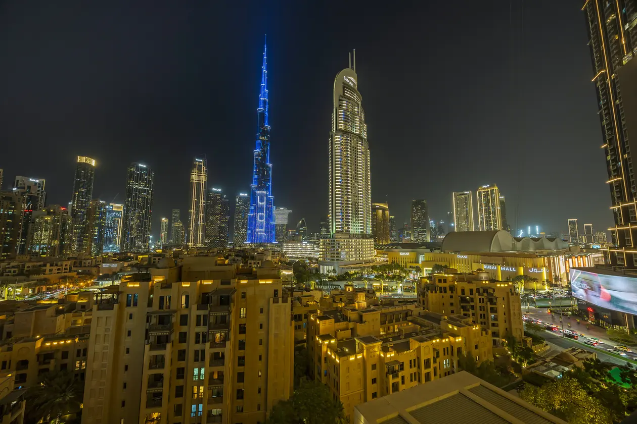 Night view of Dubai skyline at night with the blue-lit Burj Khalifa towering over a sea of illuminated skyscrapers and warm foreground buildings.