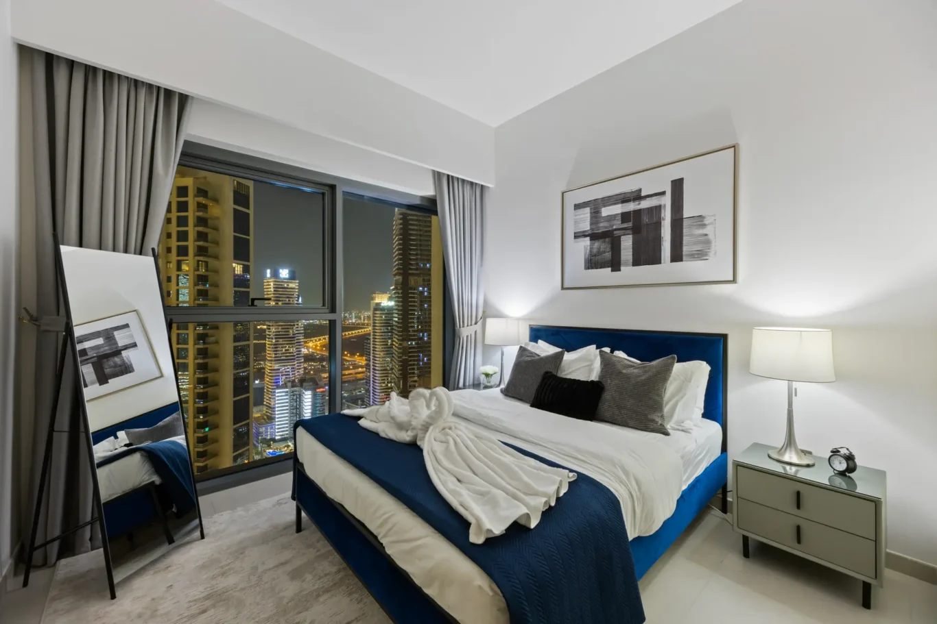 A hotel bedroom with a large window showing city lights, two blue upholstered beds, white bedding, and gray decor.