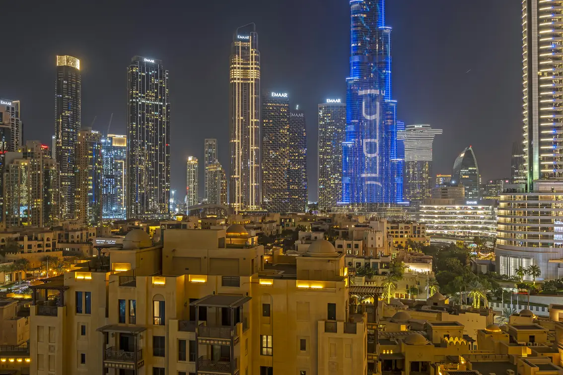 Nighttime city skyline with tall, illuminated skyscrapers and golden-lit low-rise buildings in the foreground. A nighttime view of a futuristic skyline: shimmering high-rises above a warm residential area, with a prominent blue-lit tower.