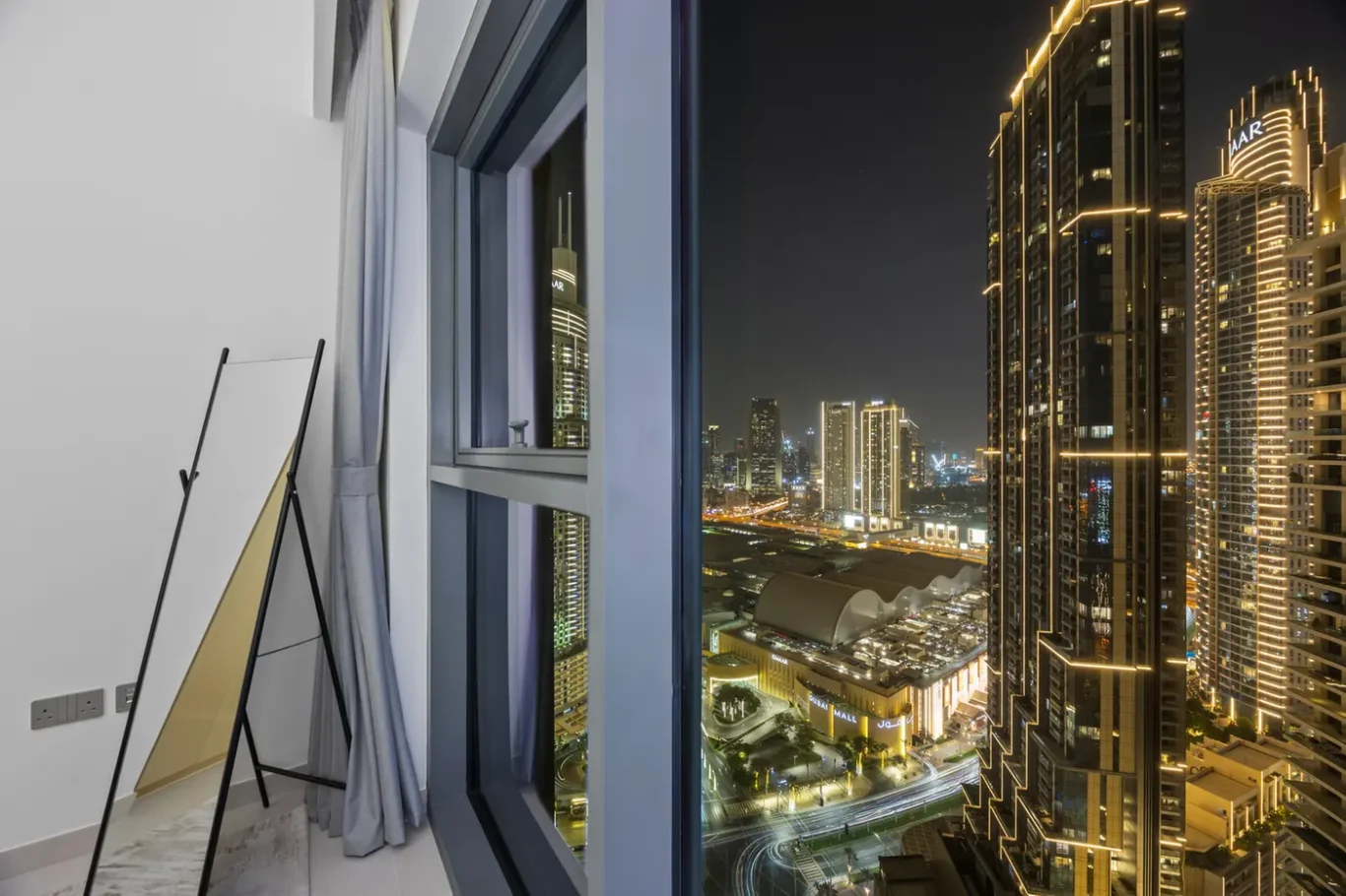 Interior of a modern room at night with a large window showing a brightly lit city skyline and tall illuminated buildings.