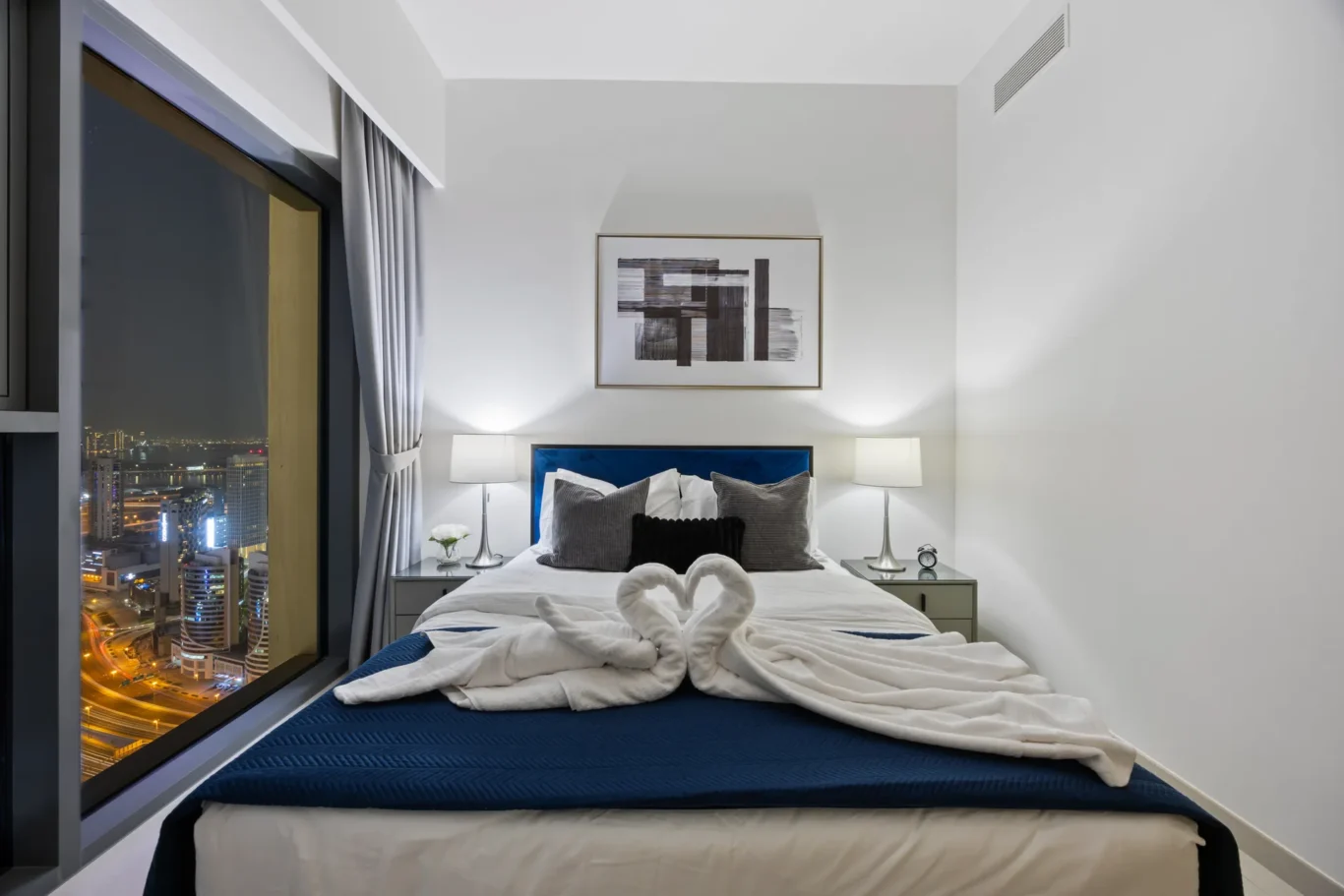 Hotel bedroom at night with a city skyline view, a white bed, blue headboard, two night lamps, and a framed abstract art piece above the bed.
