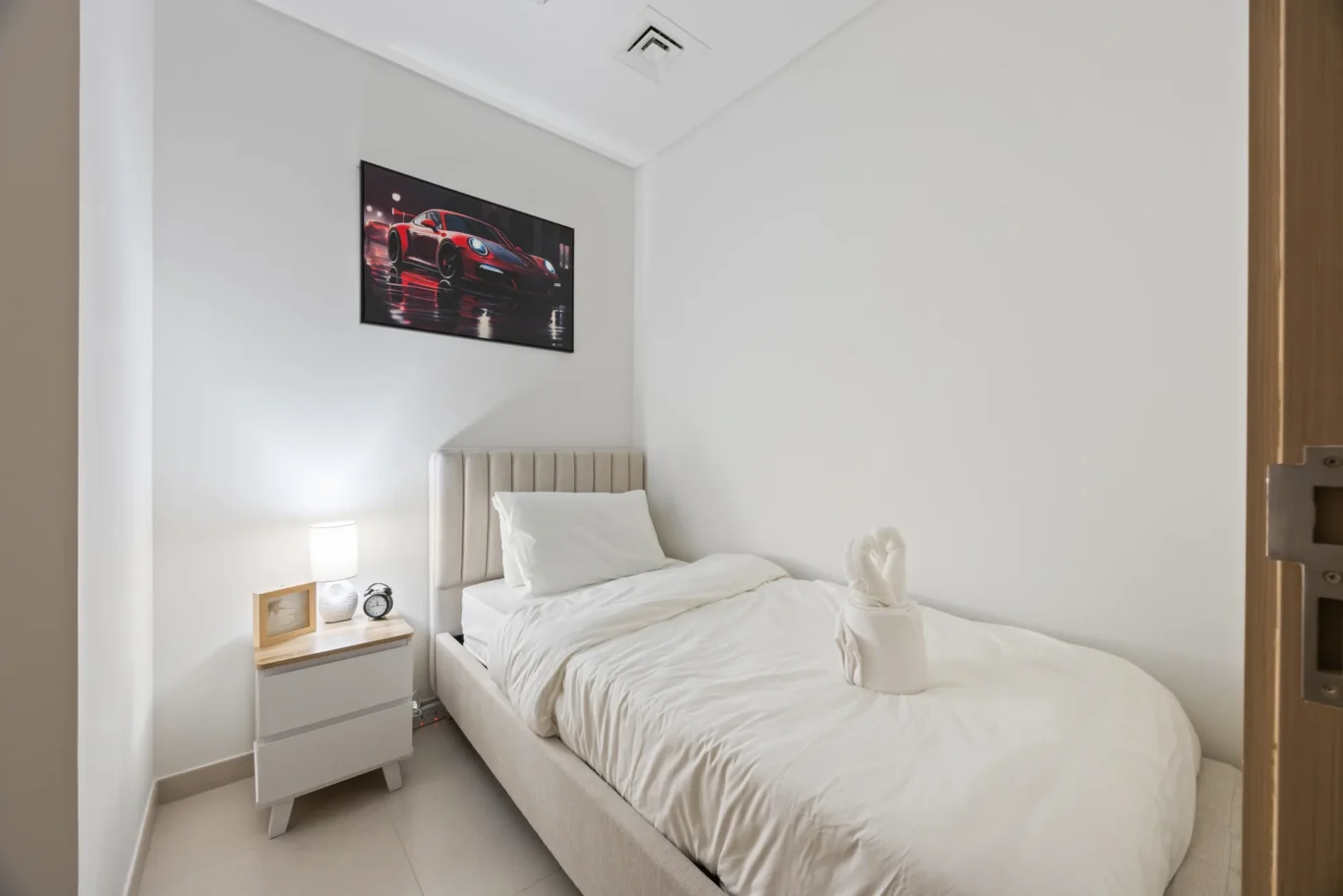 Modern, minimalist bedroom with white bedding, beige upholstered bed, a bedside table with lamp and clock, and a red sports-car wall art above the headboard.