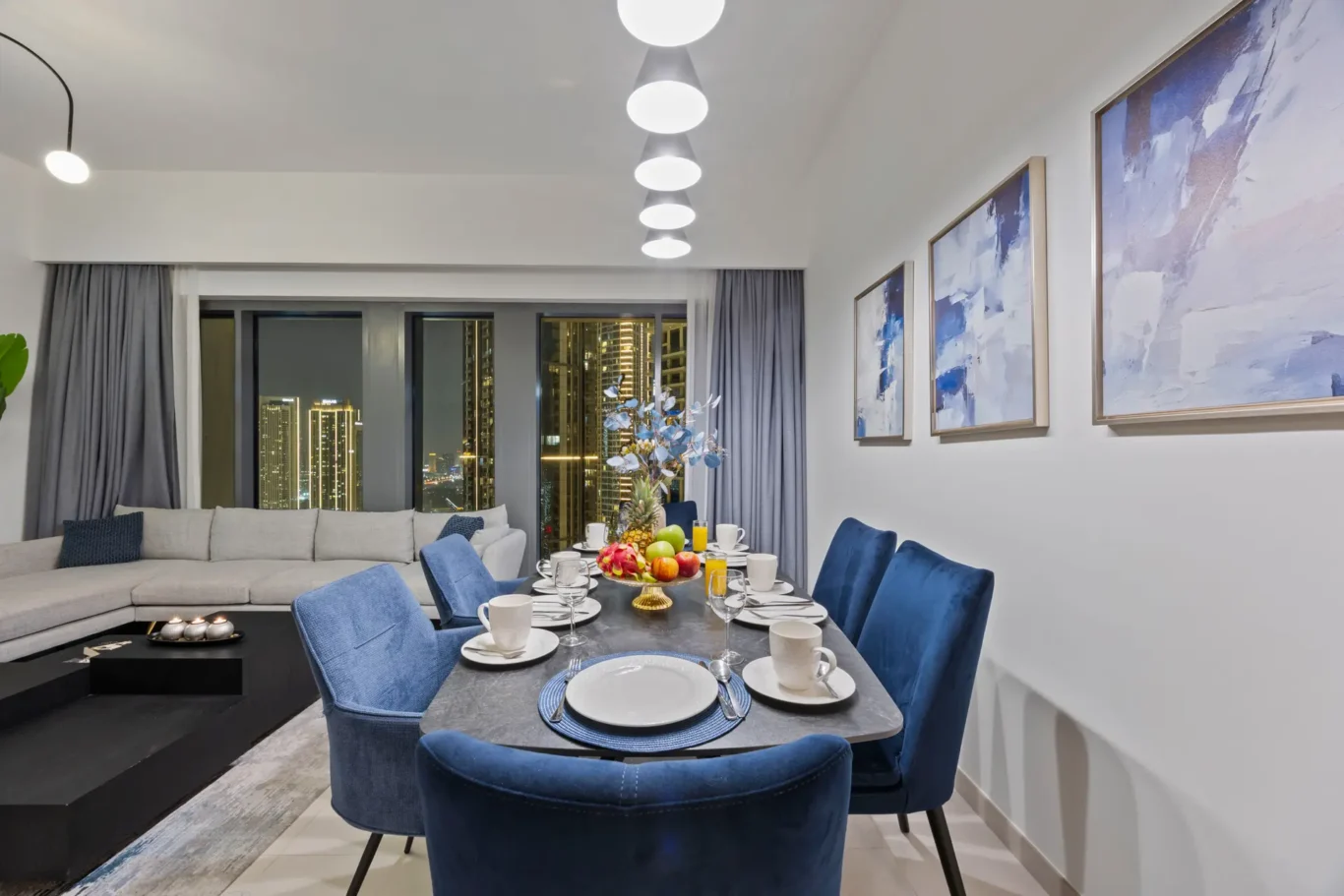 Modern dining room at night with a gray table set for eight and blue velvet chairs; city lights visible through the windows.