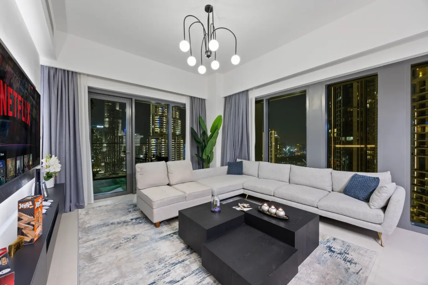 Modern living room at night with a light gray sectional sofa facing a wall-mounted Netflix screen and city skyline outside the windows.