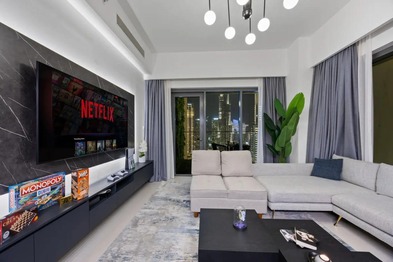 Modern living room at night with a large wall-mounted Netflix TV, grey sectional sofa, and a city skyline view through tall windows framed by blue-grey curtains.