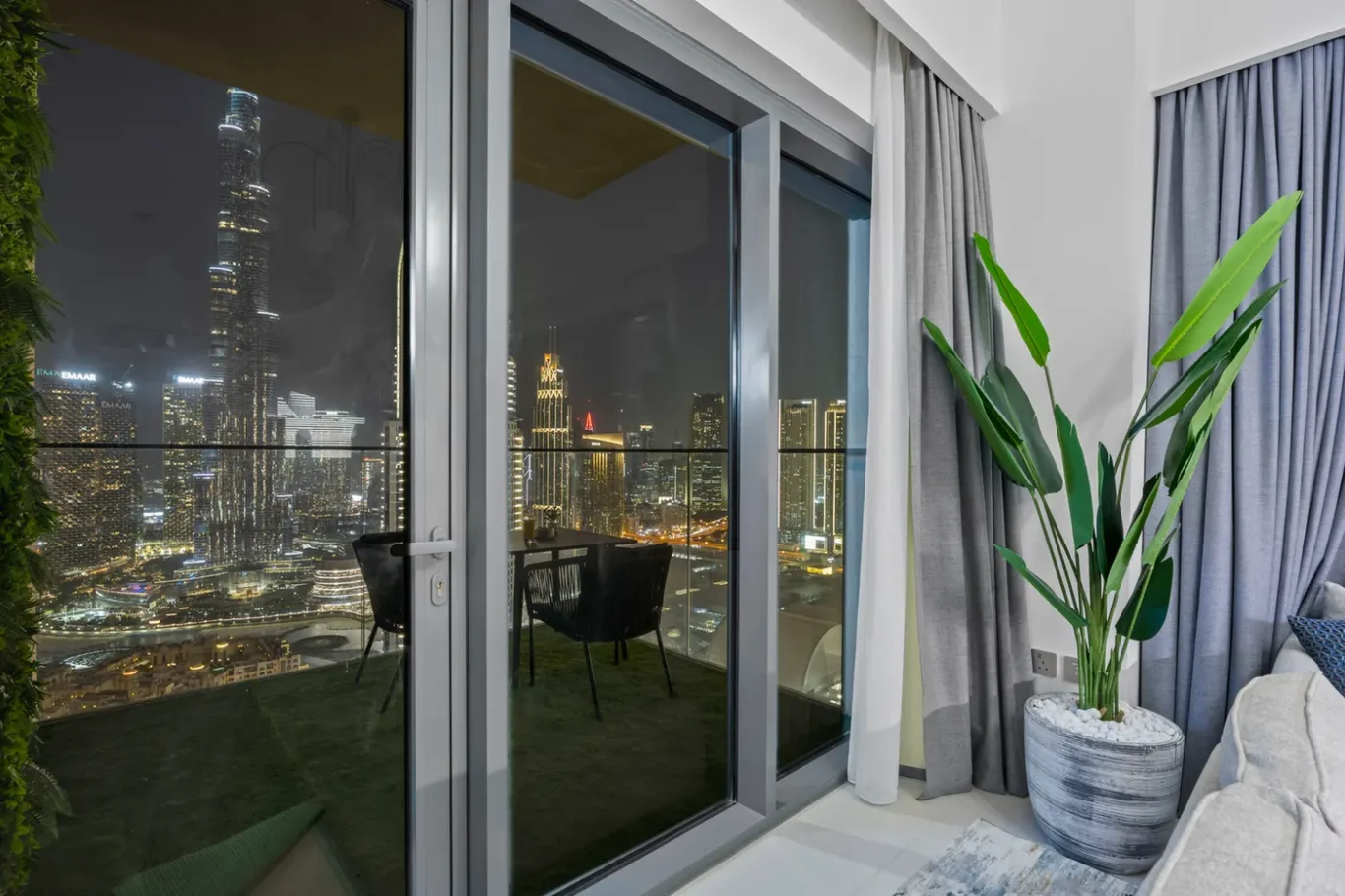 Night city skyline visible through a glass balcony door from a stylish indoor space with a large potted plant and gray curtains nearby.