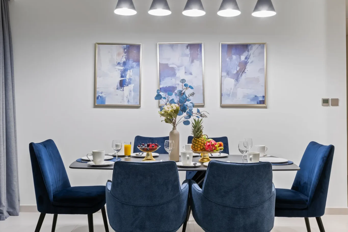 Modern dining room scene: round table set with dishes, glasses, and a central fruit and floral centerpiece, surrounded by blue upholstered chairs; three abstract blue paintings hung on the wall above the table.