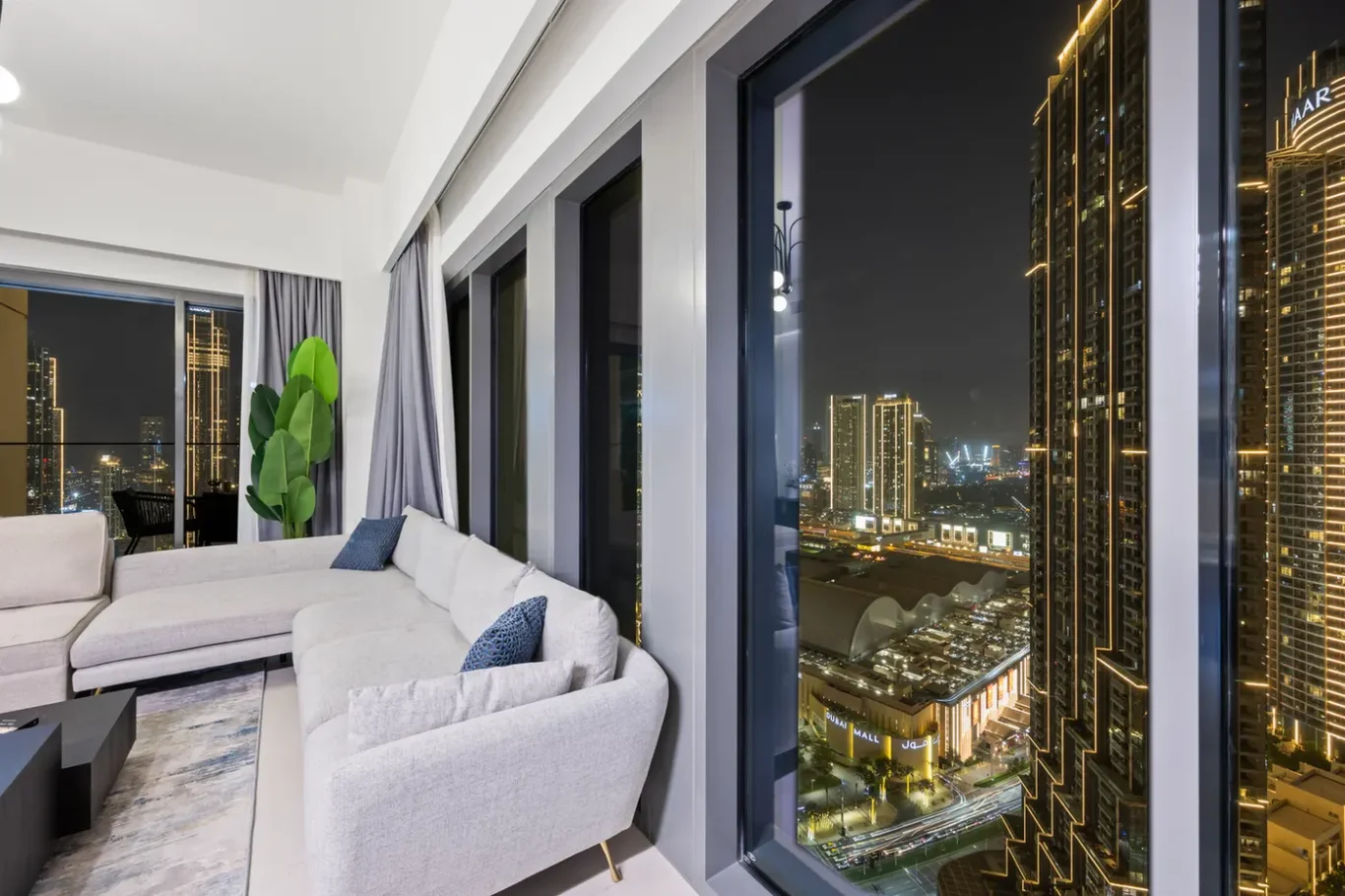 Modern living room at night with a light gray sectional sofa, blue pillows, and a tall green plant by floor-to-ceiling windows overlooking a brightly lit city skyline with high-rise buildings.